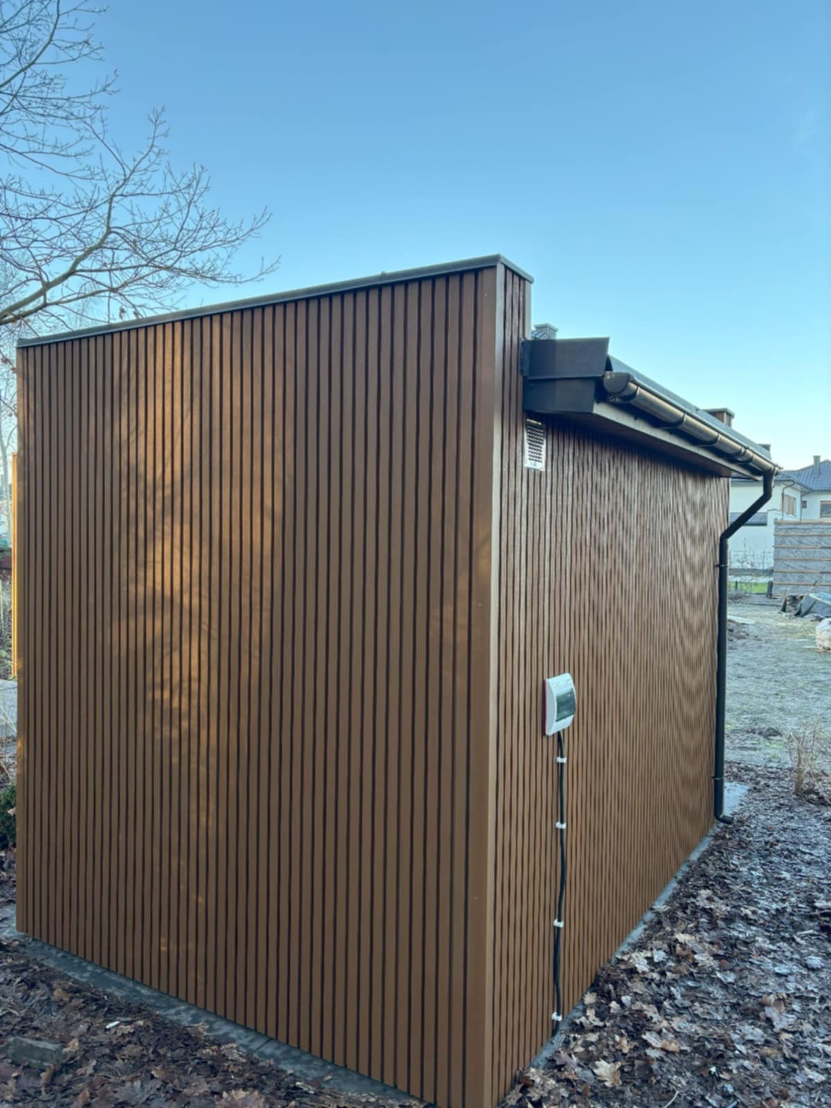 Nowoczesna sauna zewnętrzna z brązową, pionową okładziną drewnianą i czarną rynną, z zamontowaną ładowarką elektryczną na bocznej ścianie, w zimowym otoczeniu.