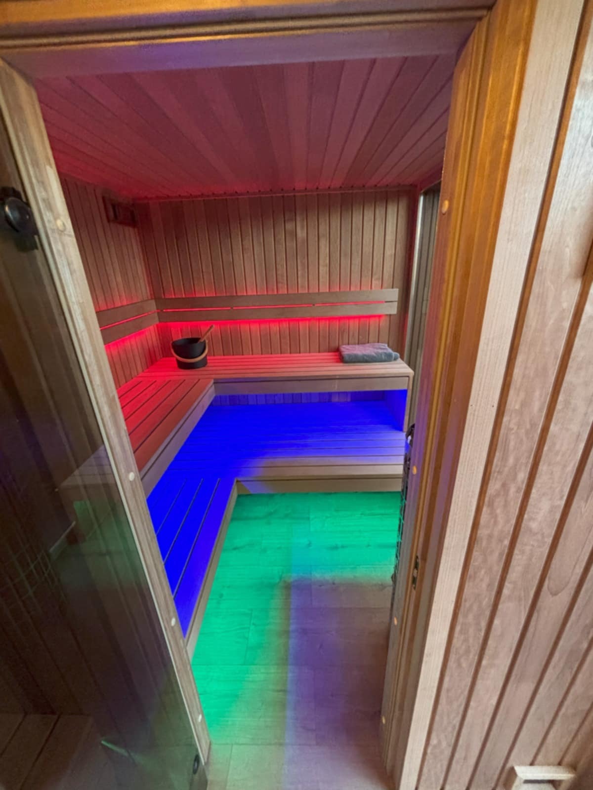 Nowoczesna sauna z drewnianym wykończeniem, podświetlona kolorowymi diodami LED: czerwonymi, niebieskimi i zielonymi. Widoczny kubeł na wodę i ręcznik na ławce.