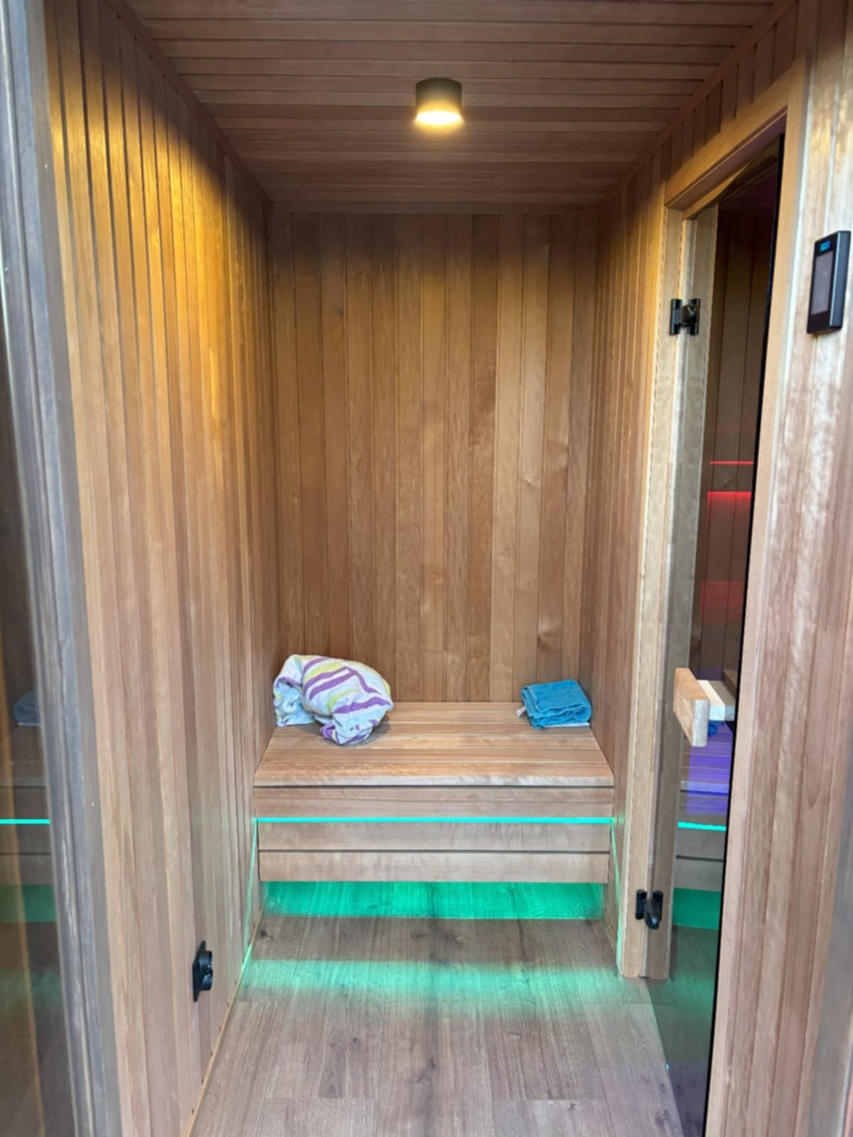 Drewniana sauna z podświetleniem LED, ręcznikami na ławce i oświetleniem sufitowym. Widok z bliska na detale wykończenia i nowoczesny design.