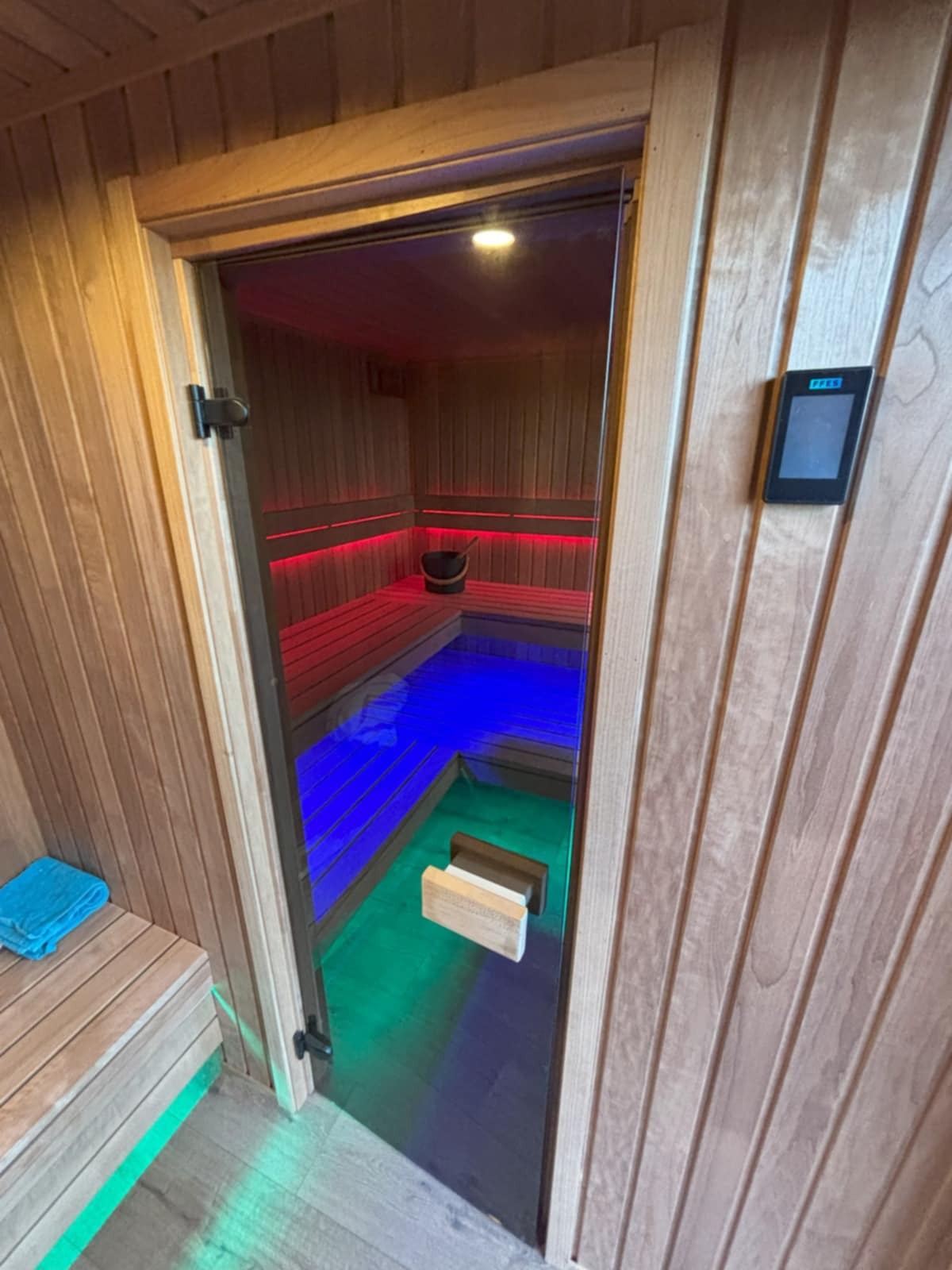 Nowoczesna sauna z drewnianym wykończeniem, podświetleniem LED w kolorach czerwonym, niebieskim i zielonym. Widoczny sterownik na ścianie i szklane drzwi.