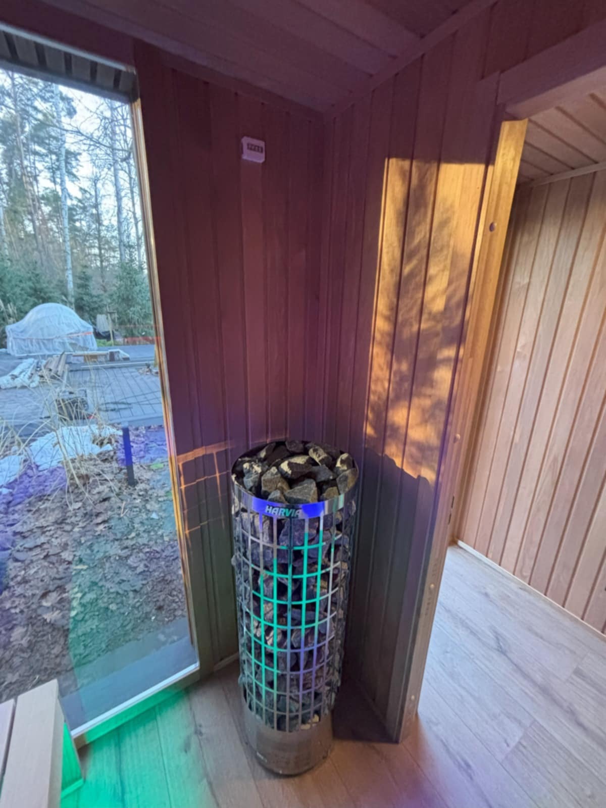 Wnętrze sauny z piecem Harvia wypełnionym kamieniami, oświetlonym na zielono i niebiesko. Widok na zewnątrz przez okno, drewniane wykończenie ścian.