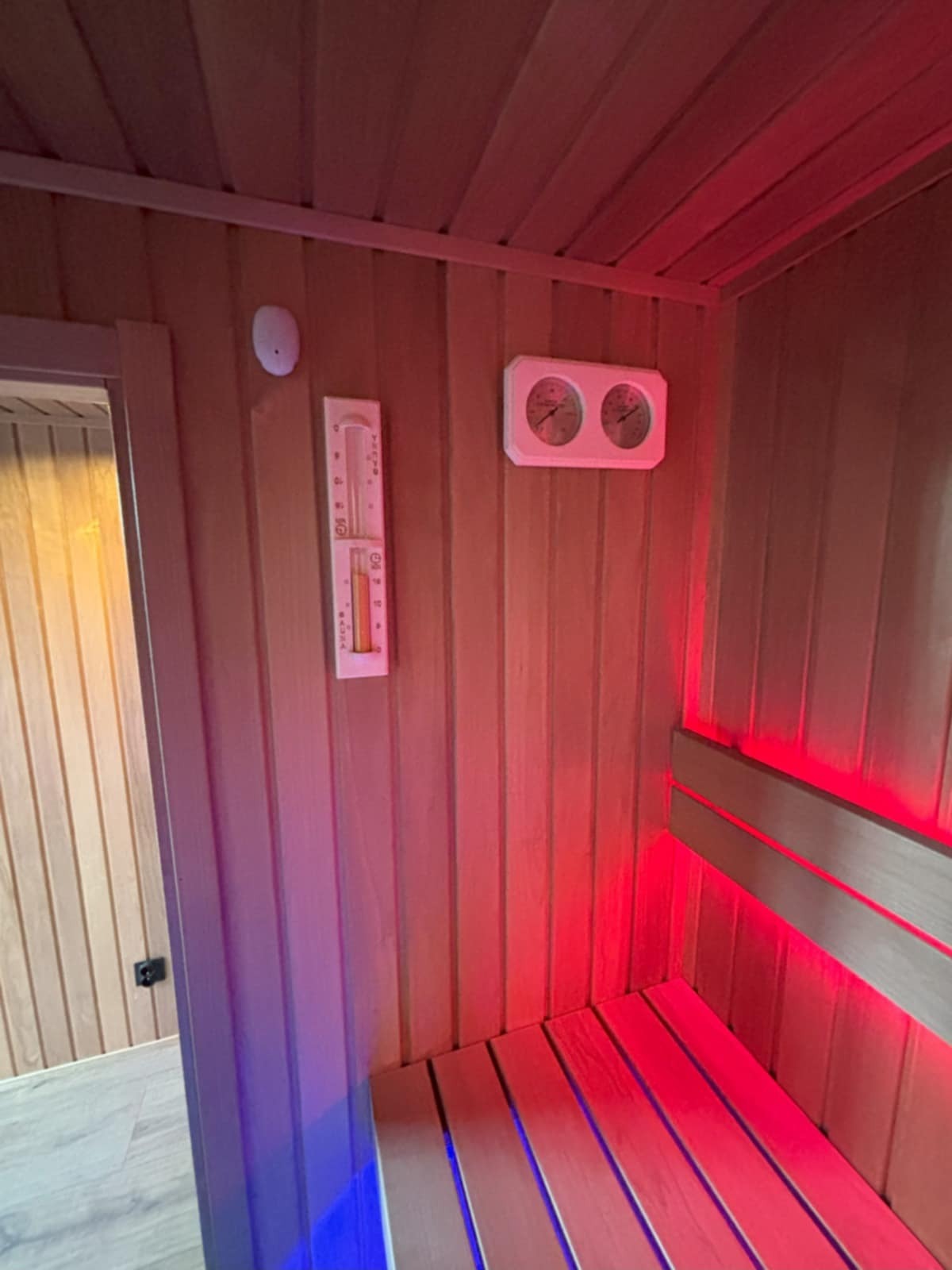 Wnętrze sauny z drewnianym wykończeniem, oświetleniem LED w kolorze czerwonym i niebieskim, termometrem oraz higrometrem na ścianie.