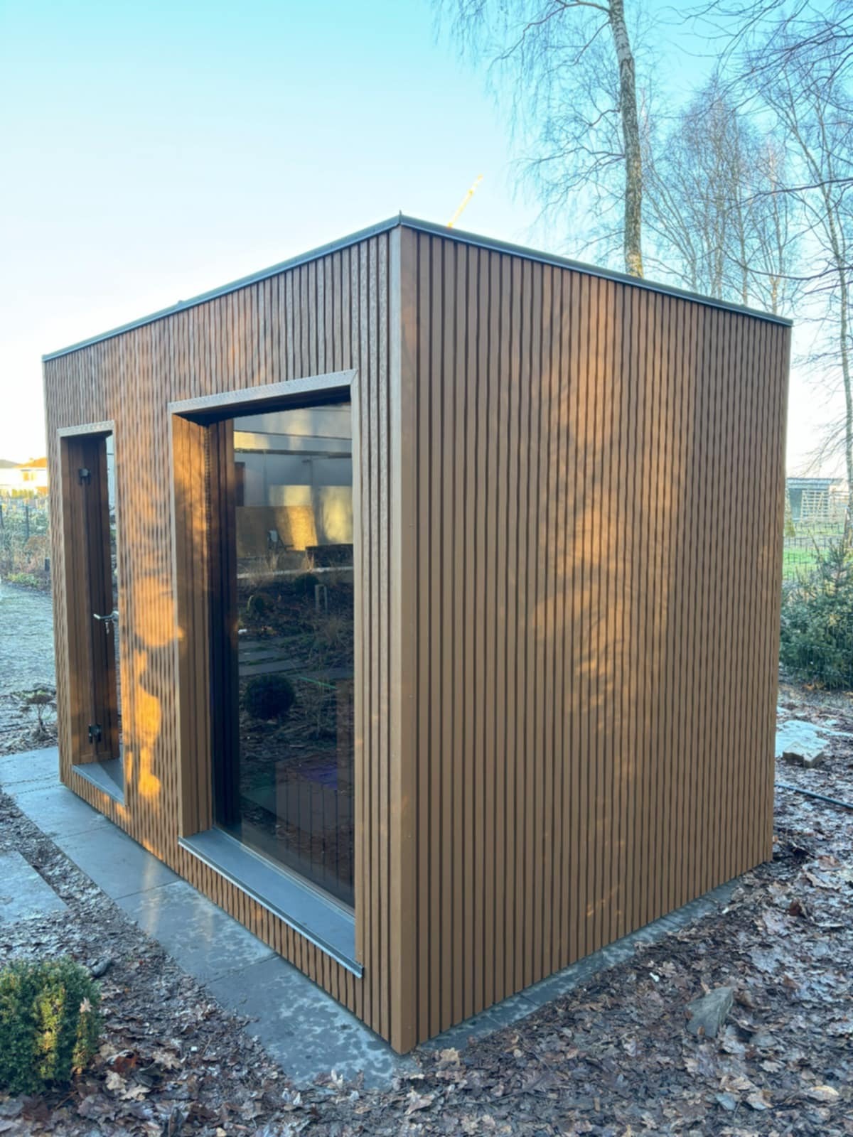 Nowoczesna, brązowa sauna modułowa z pionowymi panelami, widoczna z zewnątrz na tle ogrodu w jesiennej scenerii. Wnętrze widoczne przez okno.