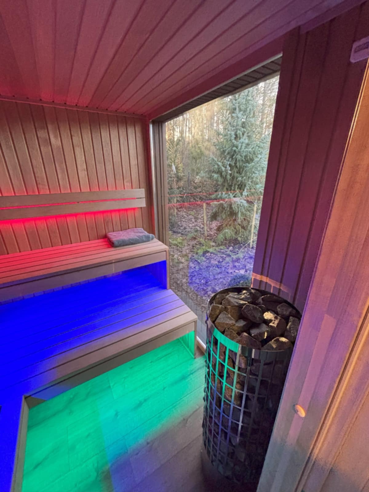 Nowoczesna sauna z oknem na zewnątrz, podświetlona kolorowymi diodami LED. Widoczne ławy, piec z kamieniami i leśny krajobraz za oknem.