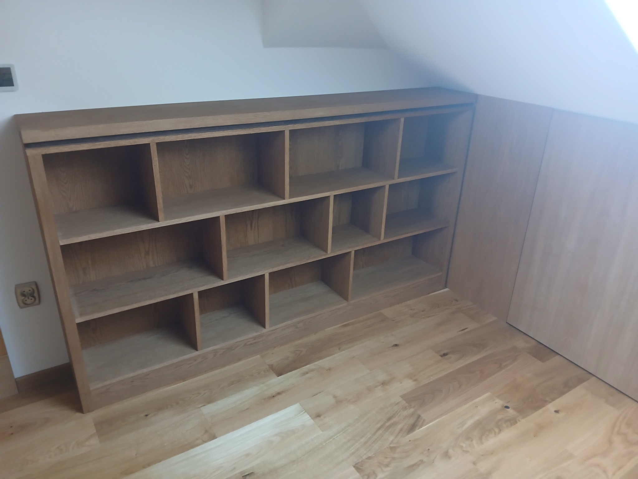 Drewniany regał na wymiar, wkomponowany w skos poddasza, z widoczną strukturą drewna i dopasowaną kolorystyką do podłogi. Widoczne gniazdko elektryczne.