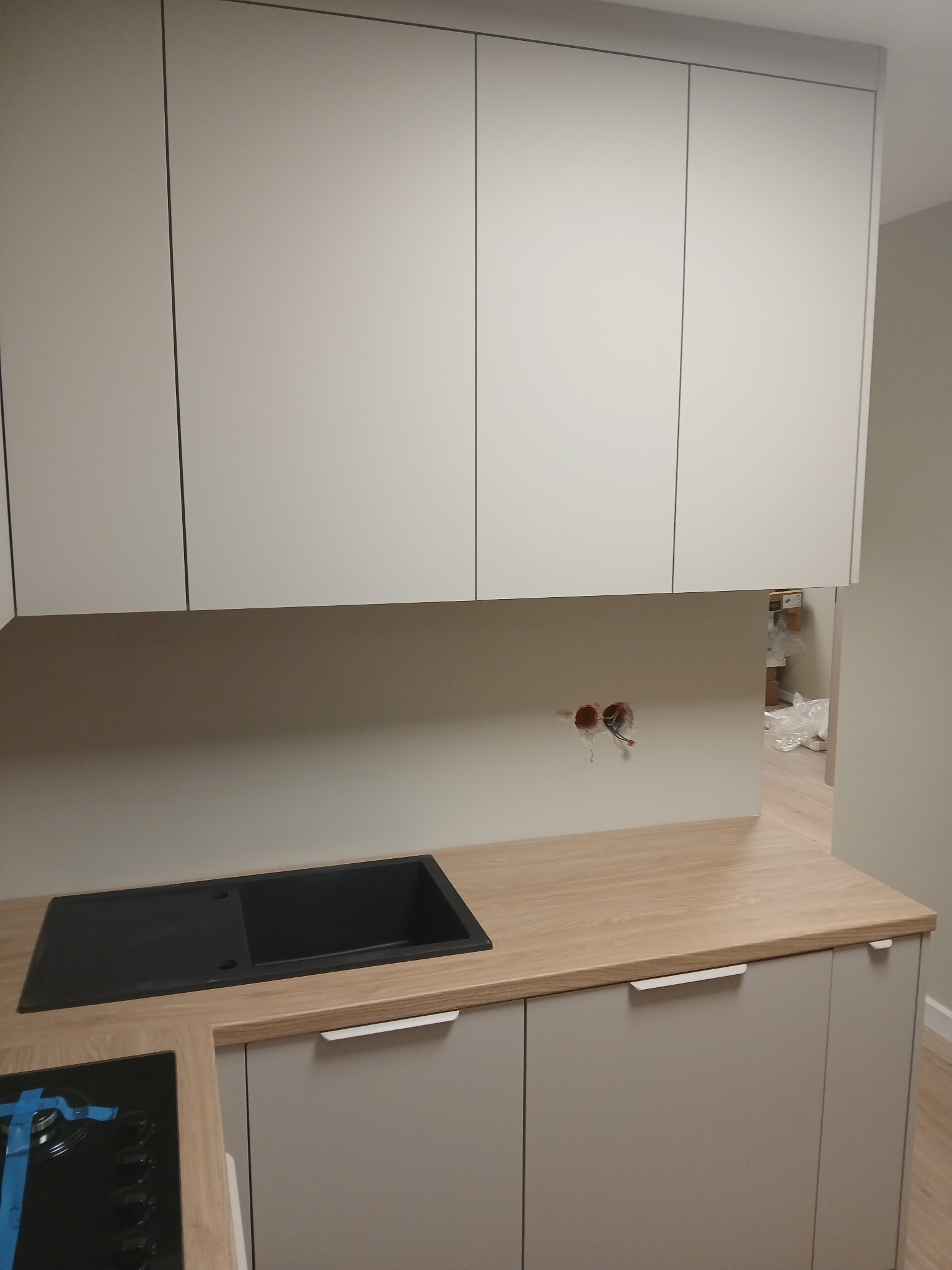 Jasna kuchnia z górnymi szafkami bez uchwytów i czarnym zlewem w drewnianym blacie. Widoczne gniazdka elektryczne i fragment płyty grzewczej.