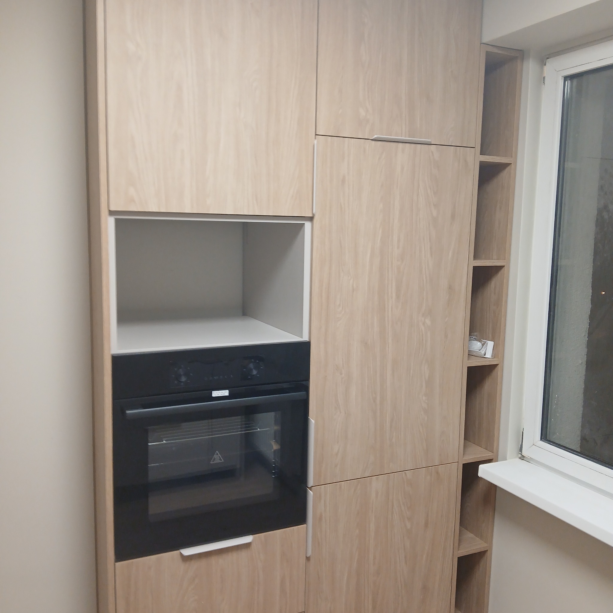 Jasna zabudowa kuchenna z piekarnikiem i otwartymi półkami obok okna. Fronty z delikatnym wzorem drewna, minimalistyczne uchwyty. Nowoczesny design.