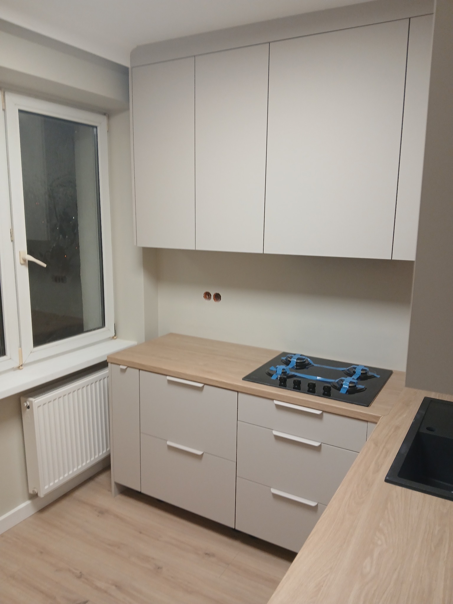 Jasna kuchnia z drewnianym blatem i płytą gazową zabezpieczoną folią. Minimalistyczne szafki wiszące i dolne z szufladami. Nowoczesny design, świeżo po montażu.