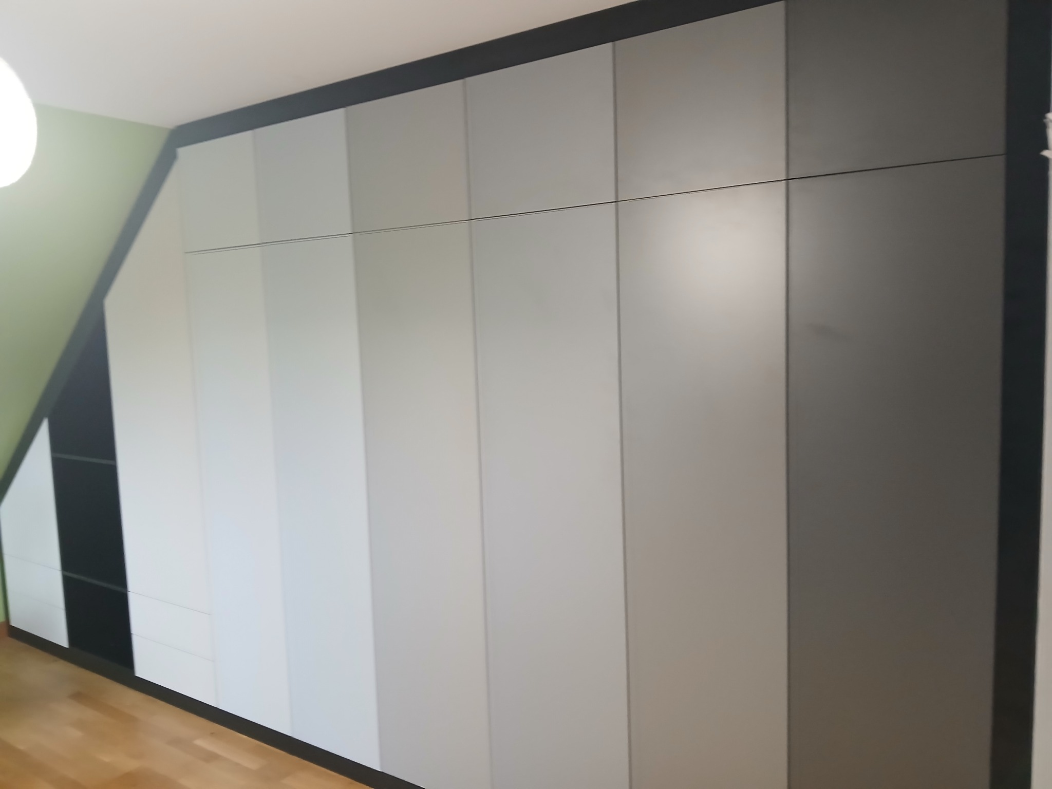 Szafa wnękowa na poddaszu ze skośnym sufitem, fronty w odcieniach szarości i czerni, minimalistyczny design, drewniana podłoga, jasne wnętrze.