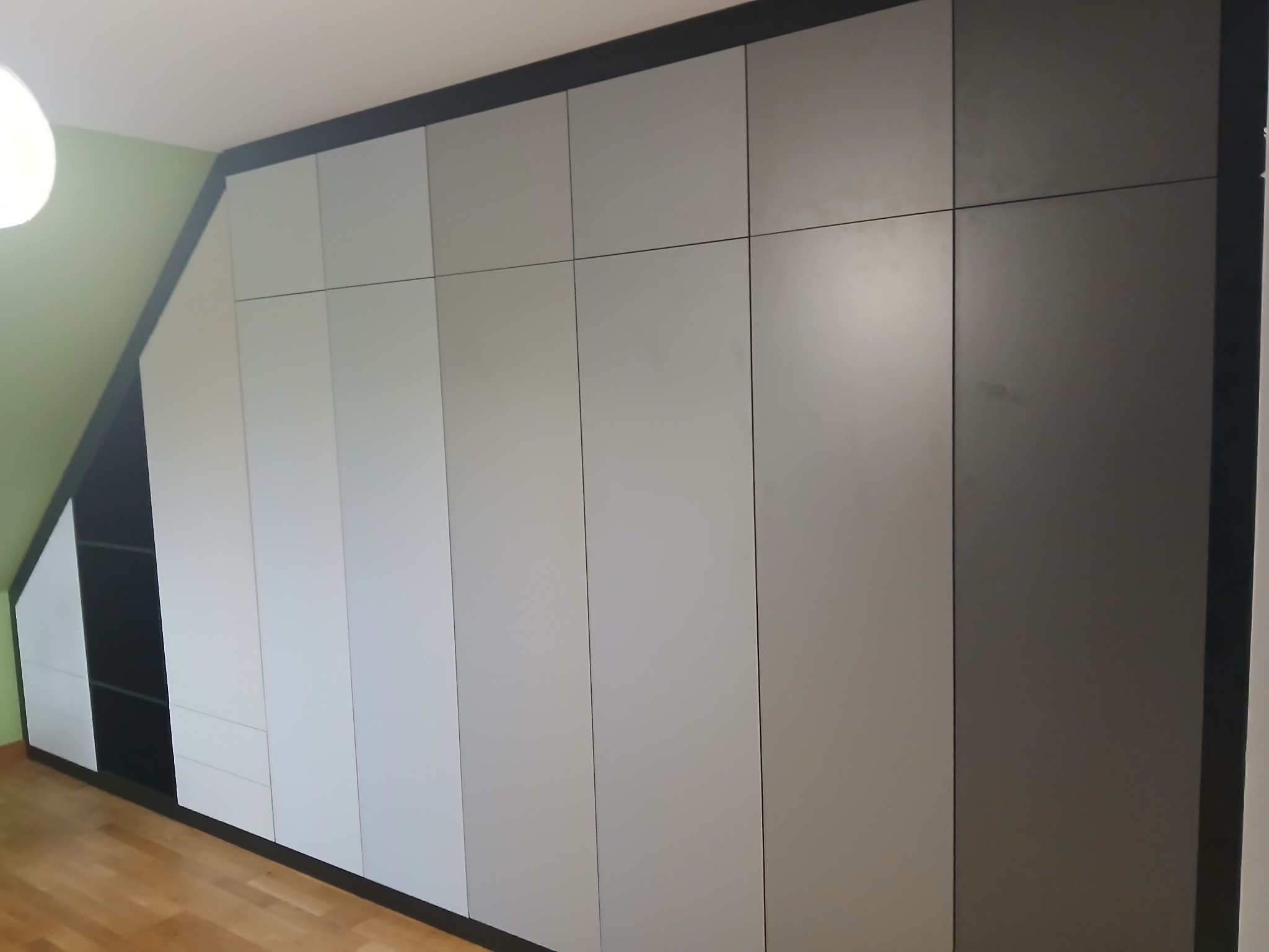 Szara szafa wnękowa z czarnymi akcentami, dopasowana do skosu poddasza, z widocznymi półkami po lewej stronie i drewnianą podłogą.