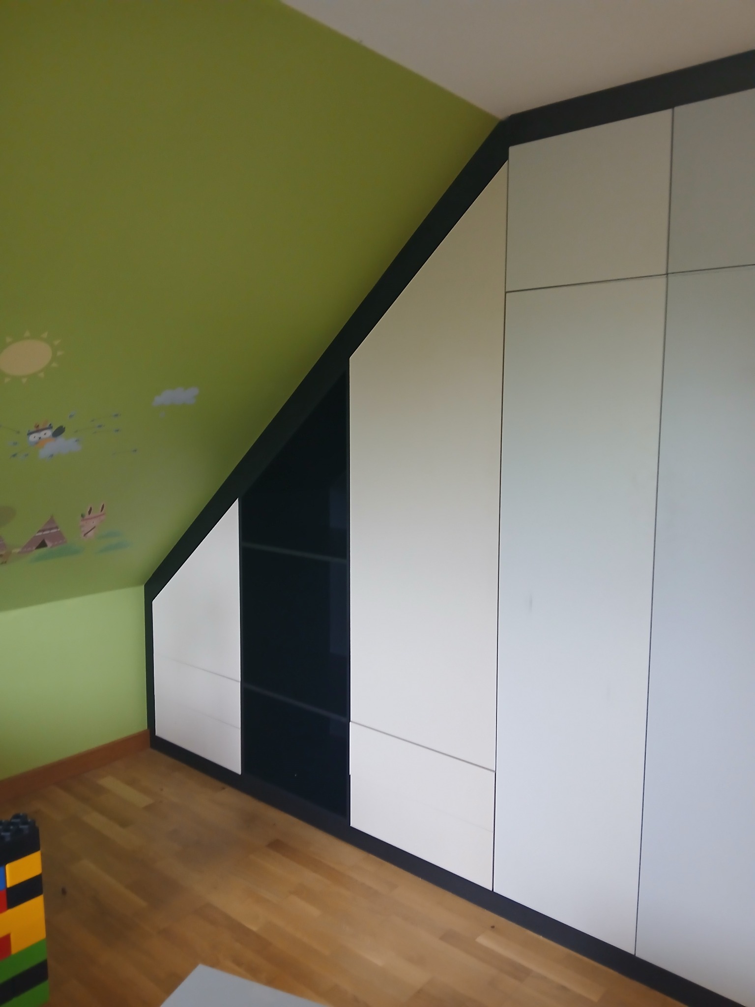 Biało-czarna szafa wnękowa na poddaszu z zieloną ścianą w dziecięce wzory. Minimalistyczny design, dopasowanie do skosu dachu, widoczny fragment podłogi.