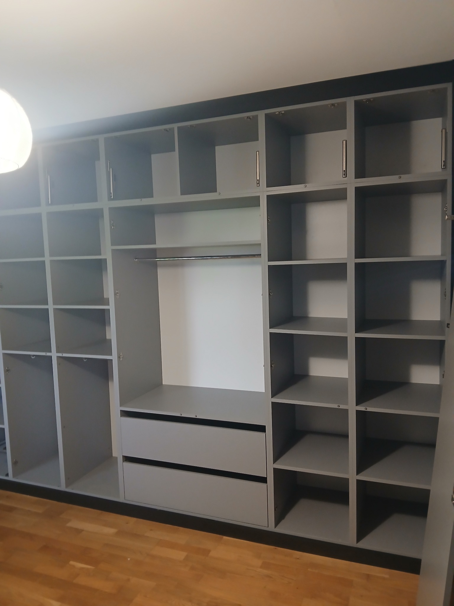 Szara szafa wnękowa z półkami i szufladami, widok z dołu, otwarte drzwi po prawej stronie, drewniana podłoga, ciemny pas na górze szafy.