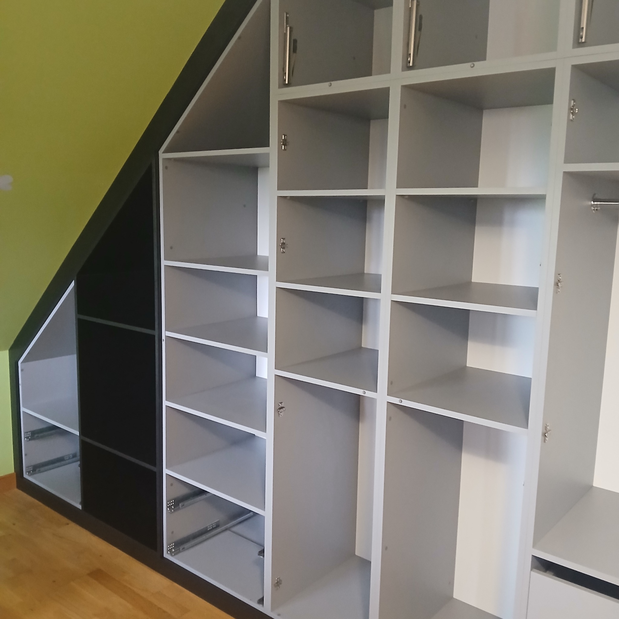 Szara szafa wnękowa z półkami i szufladami, dopasowana do skosu poddasza, z czarnym elementem po lewej stronie, na tle zielonej ściany i drewnianej podłogi.