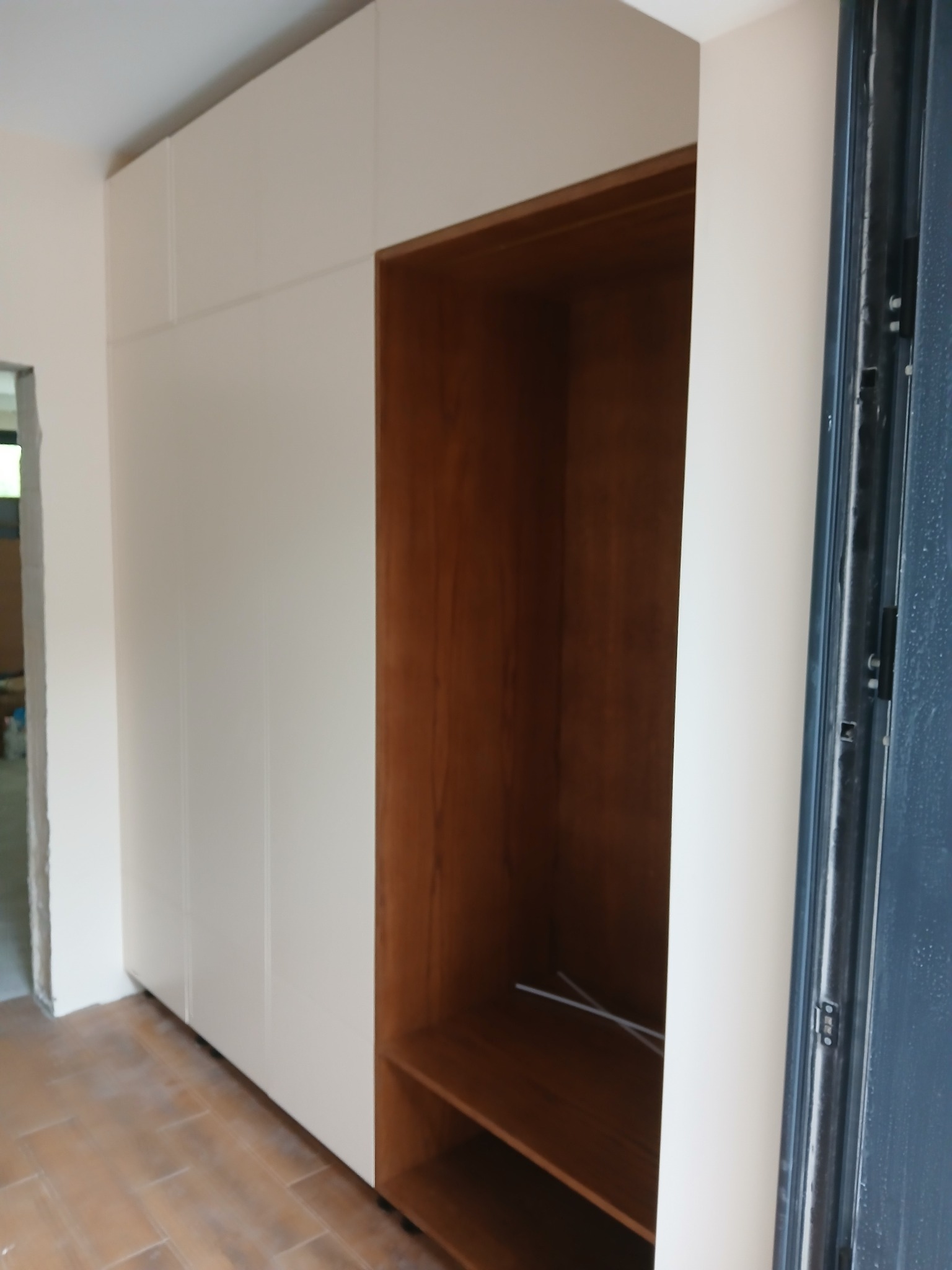 Biała szafa wnękowa z drewnianym regałem, widoczna na tle jasnego pomieszczenia z brązową podłogą. Minimalistyczny design, proste linie, wnętrze regału w ciepłym odcieniu drewna.