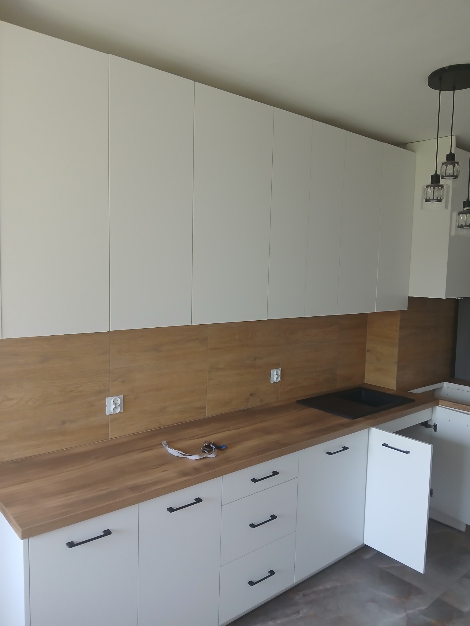 Nowoczesna kuchnia z białymi szafkami i drewnianym blatem. Uwagę przykuwają czarne, minimalistyczne uchwyty i oświetlenie. Widoczne gniazdka elektryczne na ścianie.