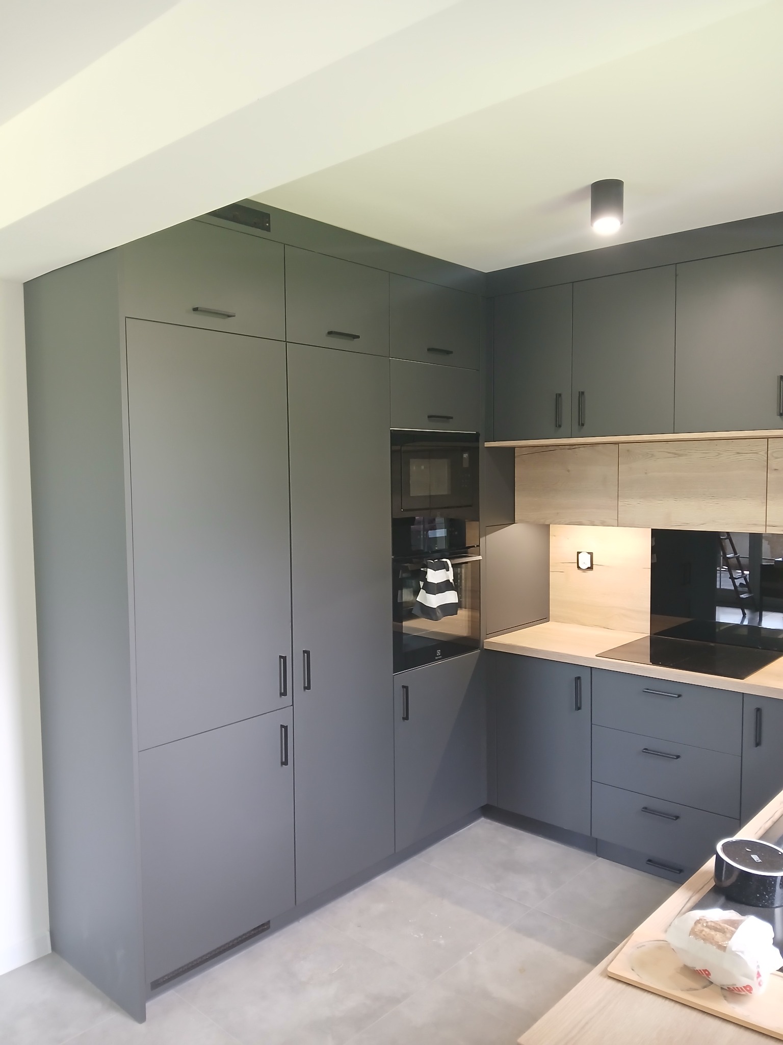 Nowoczesna kuchnia w odcieniach szarości z drewnianymi akcentami. Minimalistyczne fronty szafek, zabudowane sprzęty AGD i subtelne oświetlenie punktowe.