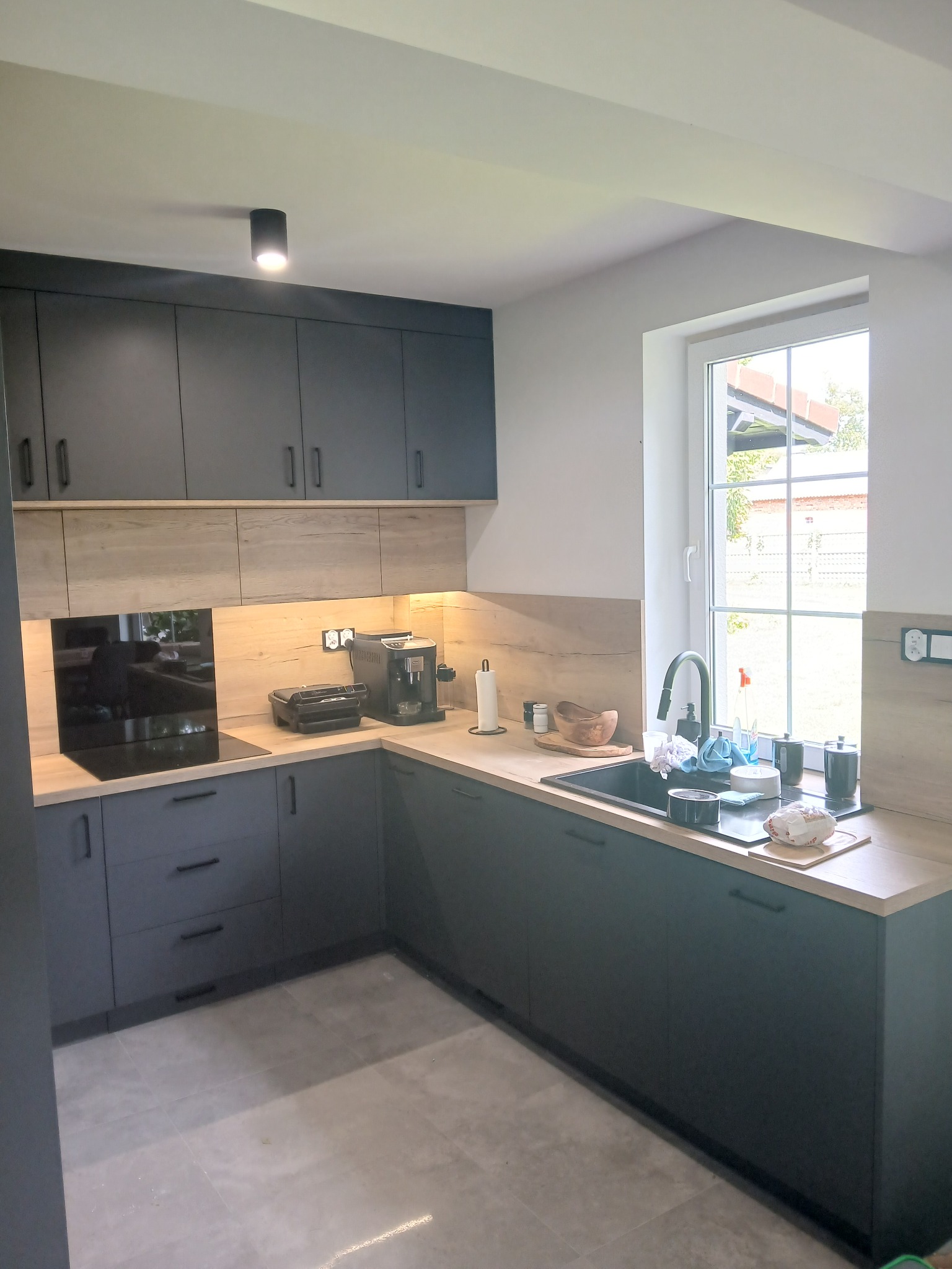 Nowoczesna kuchnia w odcieniach szarości z drewnianymi akcentami. Uwagę przykuwa czarny zlew pod oknem i minimalistyczne oświetlenie sufitowe.