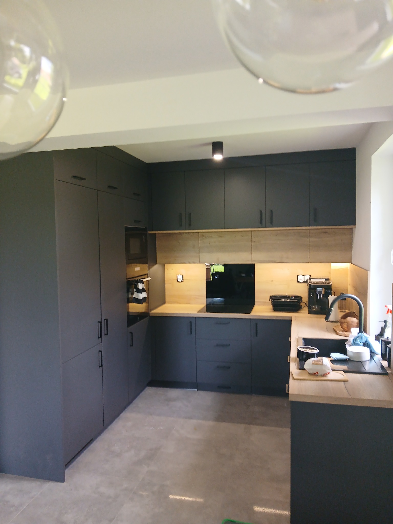 Nowoczesna kuchnia w odcieniach szarości z drewnianymi akcentami, oświetleniem punktowym i dużymi szafkami, z widocznym wyposażeniem i elementami dekoracyjnymi.