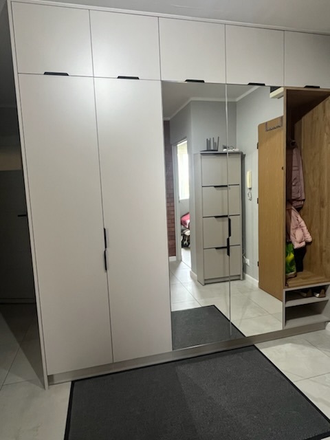 Jasna szafa wnękowa z lustrem i czarnymi uchwytami, zintegrowana z wieszakiem na ubrania. Widoczny fragment przedpokoju z szarą wycieraczką.