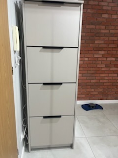 Wysoka, wąska szafka na buty z czterema szufladami w odcieniach szarości, z czarnymi uchwytami, na tle ściany z czerwonej cegły i jasnej podłogi.