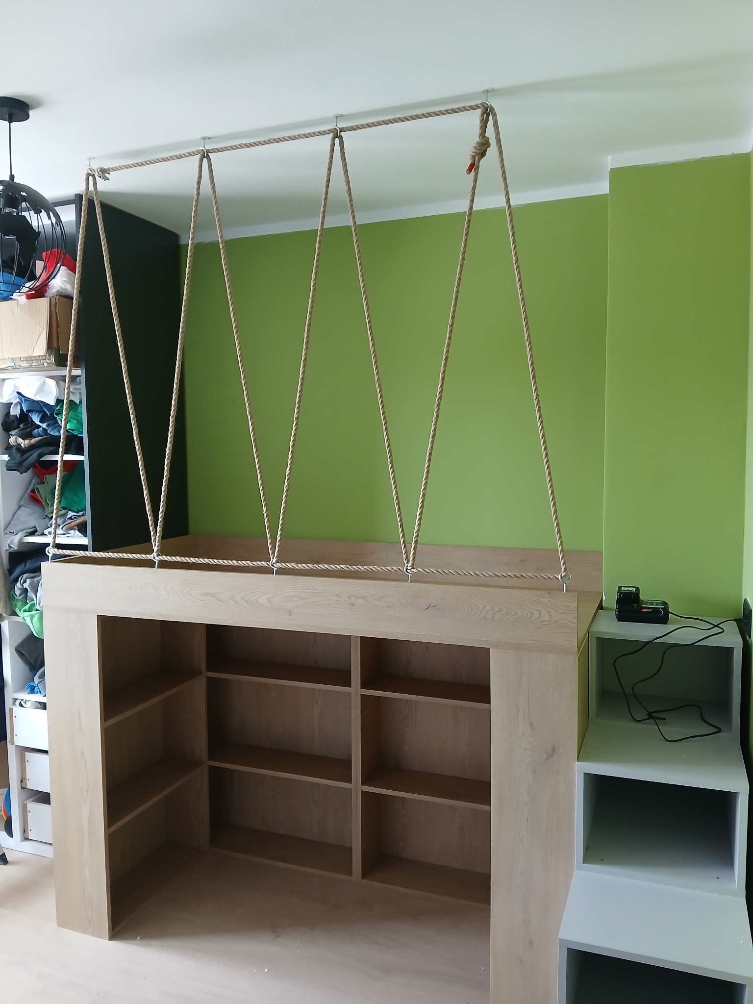 Kreatywna konstrukcja z jasnego drewna z półkami, zawieszona na linach przymocowanych do sufitu, na tle zielonej ściany. Nietypowy mebel w nowoczesnym wnętrzu.