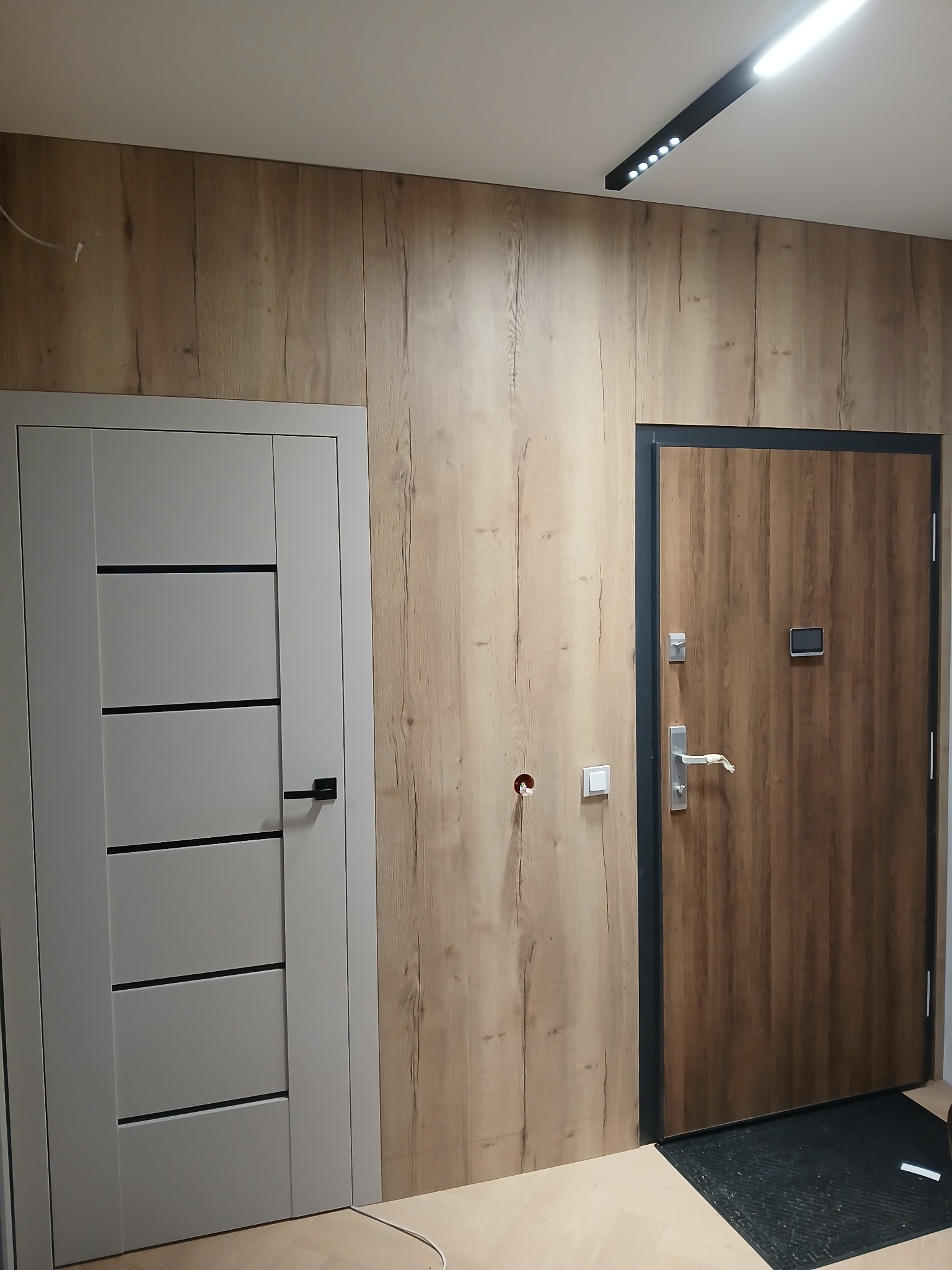 Nowoczesny przedpokój z dwiema parami drzwi: wewnętrznymi szarymi i zewnętrznymi w okleinie drewnopodobnej z czarną ramą. Ściana wykończona panelami drewnopodobnymi.