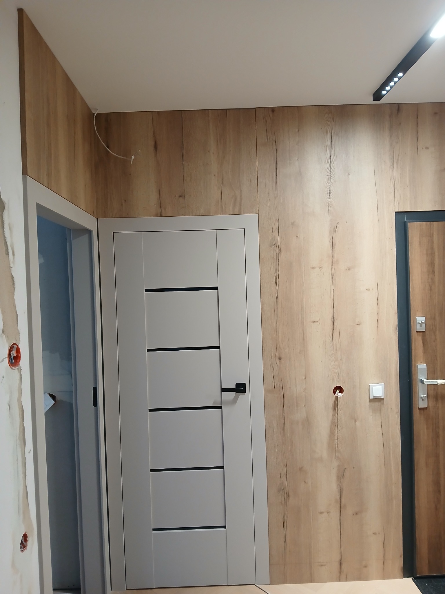 Wykończone wnętrze z panelami ściennymi imitującymi drewno, szarą, nowoczesną stolarką drzwiową i oświetleniem punktowym. Widoczne gniazdka elektryczne.