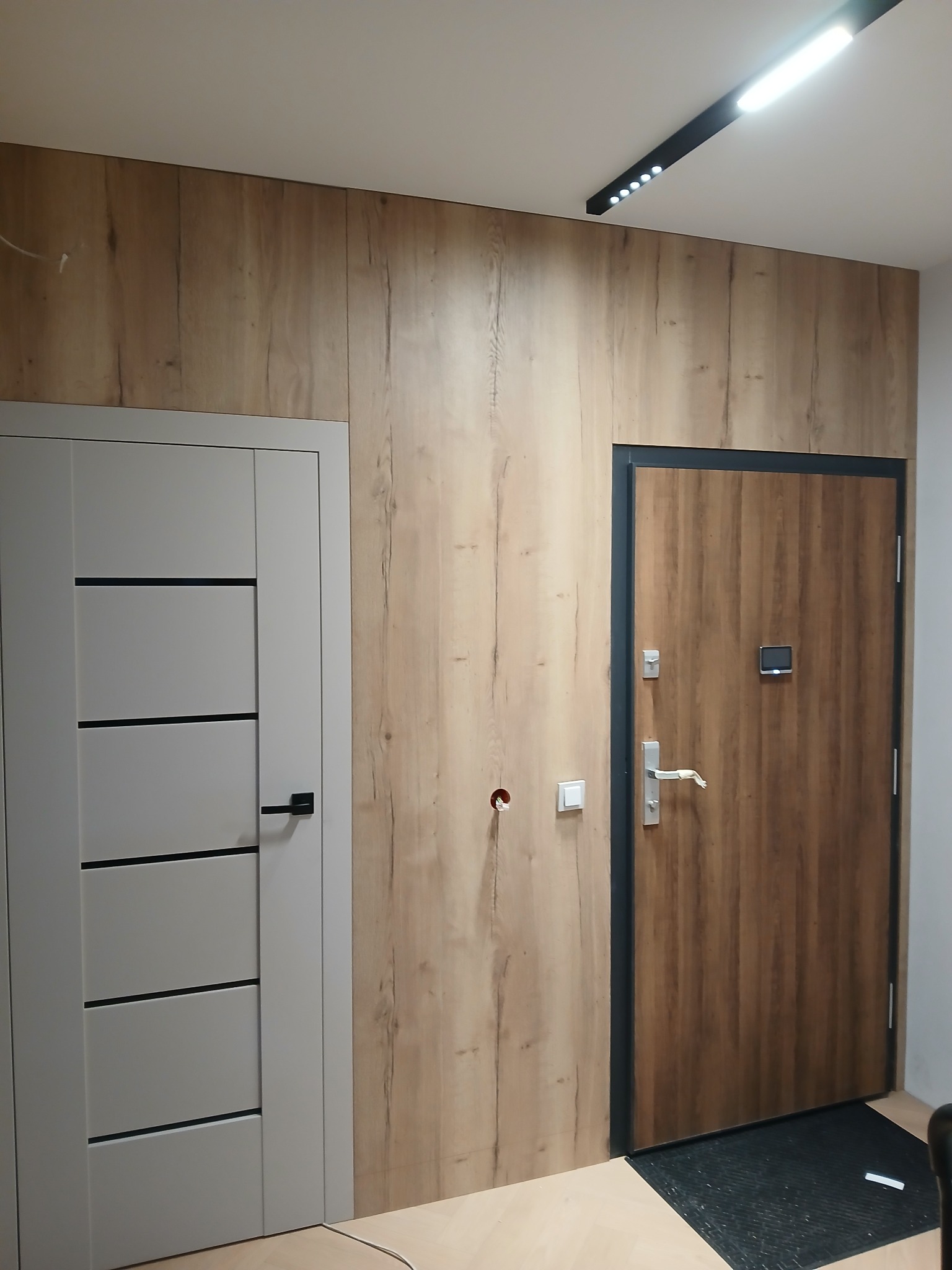 Nowoczesny przedpokój z drewnianą okładziną ścienną, dwojgiem drzwi (białe i brązowe) oraz oświetleniem LED. Widoczne gniazdka elektryczne.