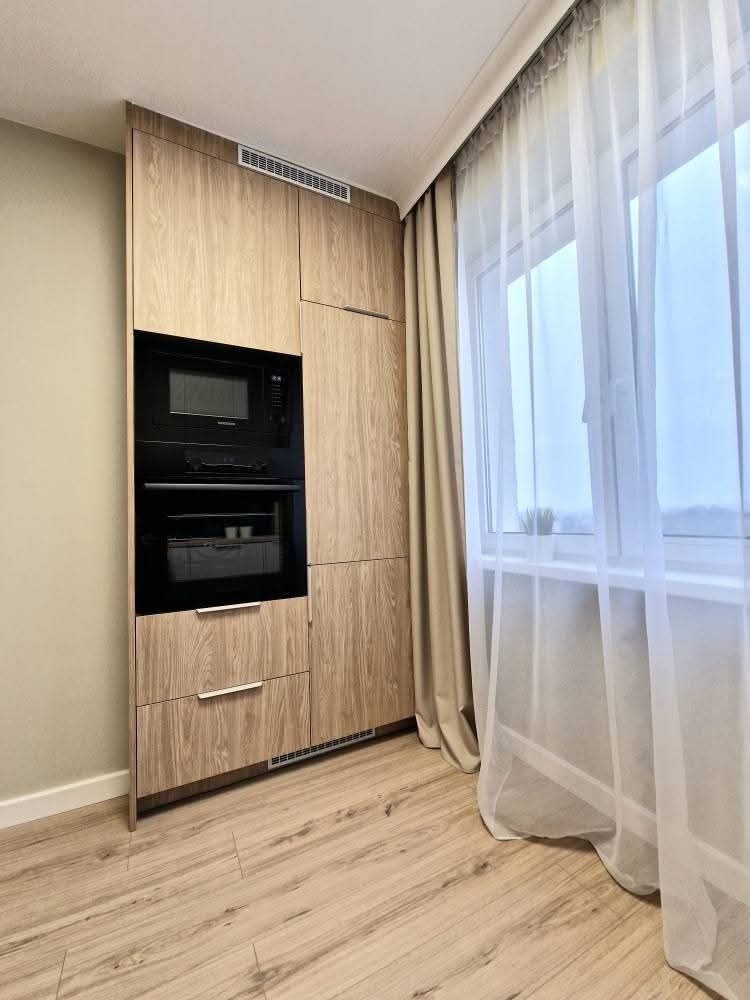 Drewniana zabudowa kuchenna z piekarnikiem i mikrofalówką, zintegrowana z szafkami, na tle okna z delikatnymi firanami i drewnianej podłogi.