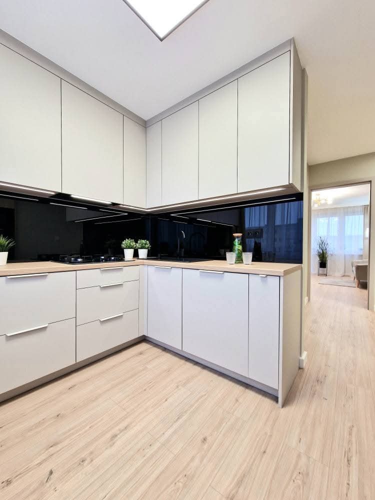 Jasna kuchnia w kształcie litery L z drewnianym blatem i czarnym panelem nad blatem. Minimalistyczny design, oświetlenie LED i drewniana podłoga.