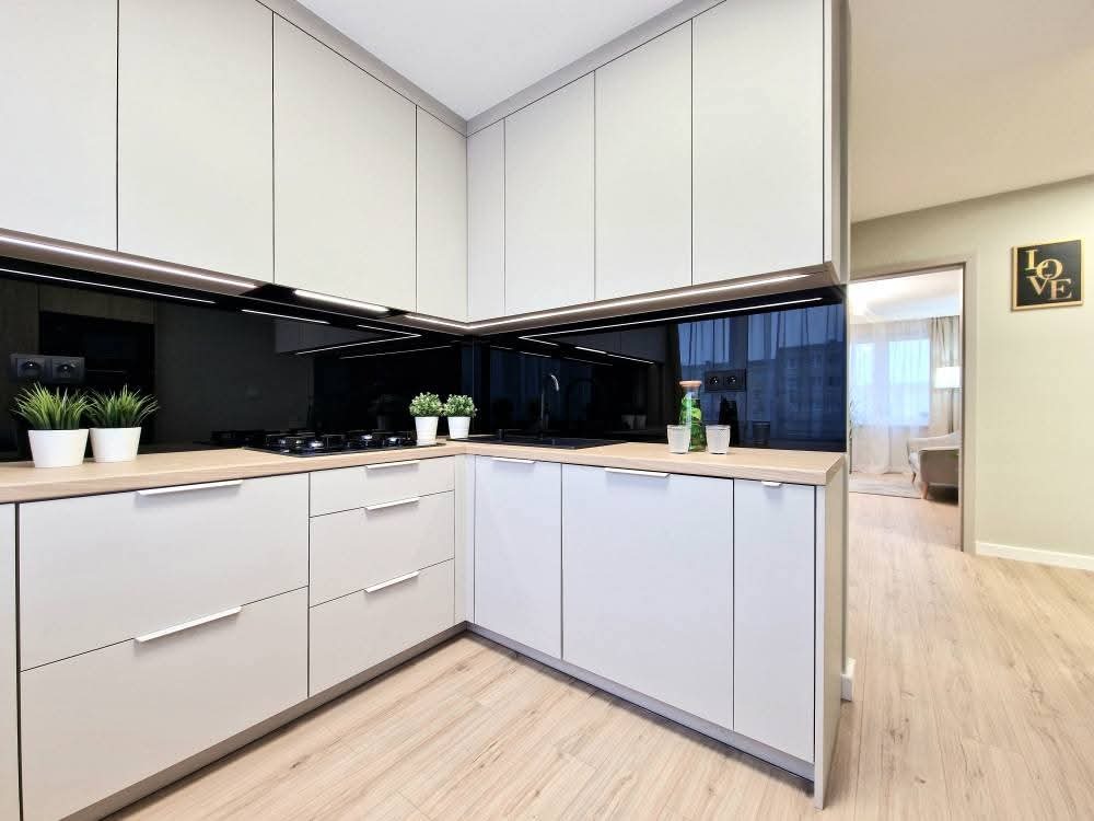 Jasna kuchnia na wymiar z czarnym panelem szklanym, oświetleniem LED i drewnianym blatem. Widok na salon z drewnianą podłogą i dekoracją ścienną.