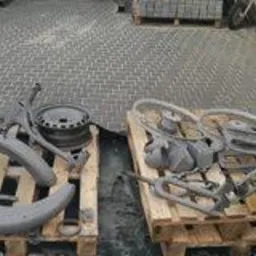 Metalowe elementy motocyklowe, w tym błotniki, rama i felgi, ułożone na paletach. Części pokryte szarą, matową powłoką, prawdopodobnie po procesie przygotowania do malowania.