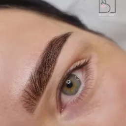 Zbliżenie na oko i brwi po stylizacji henną pudrową, widoczny precyzyjny kształt i kolor brwi oraz logo salonu w tle.