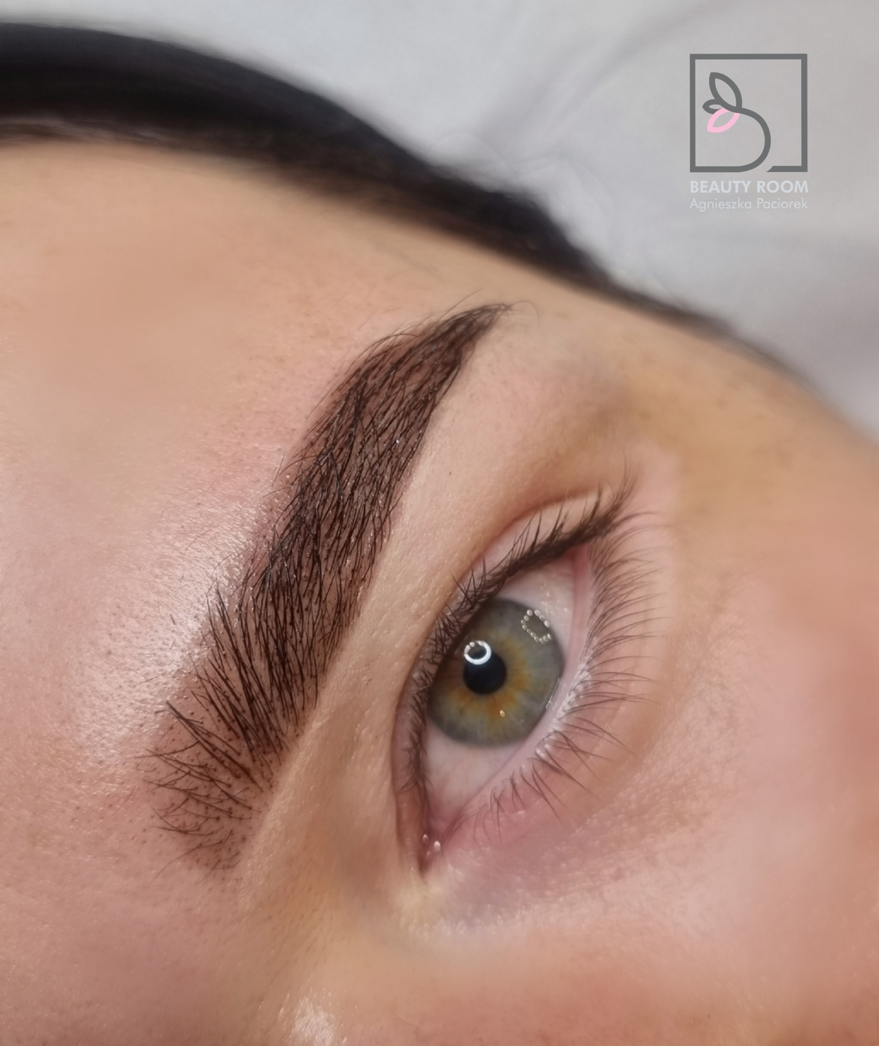 Zbliżenie na oko i brwi po stylizacji henną pudrową, widoczny precyzyjny kształt i kolor brwi oraz logo salonu w tle.