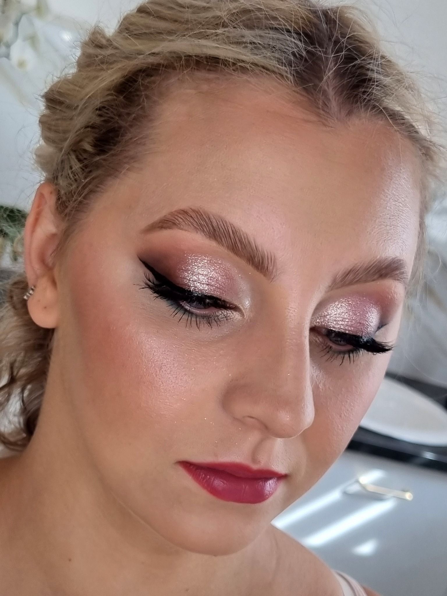 Portret kobiety z profesjonalnym, błyszczącym makijażem oczu w odcieniach różu i wyraźną kreską oraz bordowymi ustami, włosy upięte w kok