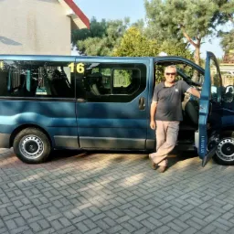 Niebieski bus z numerem '16' na szybie, kierowca w okularach opiera się o otwarte drzwi na brukowanym podjeździe.