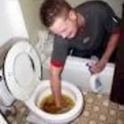 Mężczyzna z zanurzoną ręką w muszli klozetowej wypełnionej brudną wodą, próbuje usunąć zator. Widoczny fragment łazienki z beżowymi płytkami podłogowymi.