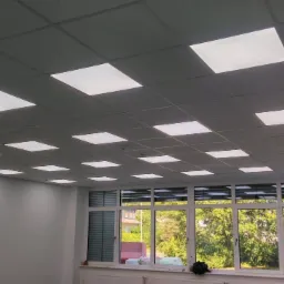 Sufit kasetonowy z równomiernie rozmieszczonymi panelami LED, widok z wnętrza pomieszczenia z dużymi oknami i widokiem na zieleń.