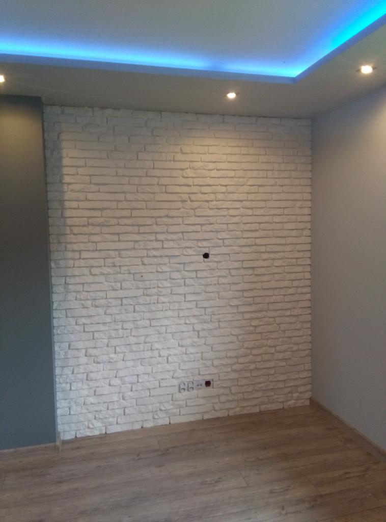 Ściana z białej cegły dekoracyjnej z widocznym gniazdkiem elektrycznym i otworem na kable, oświetlona niebieskim światłem LED na suficie.