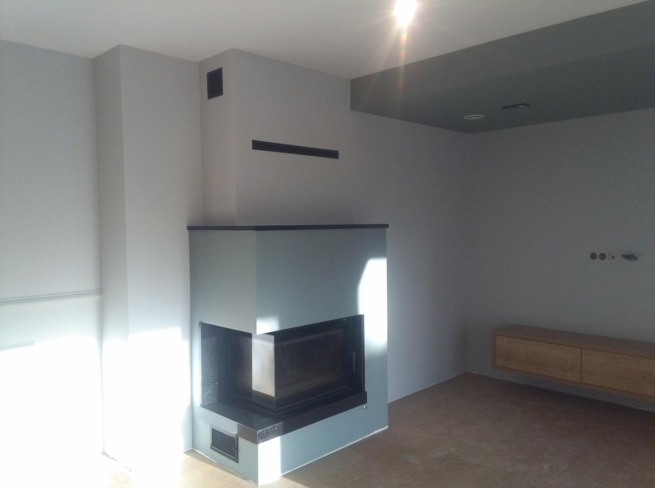 Nowoczesny narożny kominek z czarnym obramowaniem i szarymi ścianami w minimalistycznym salonie, z widoczną szafką RTV w tle i wnękami wentylacyjnymi w ścianie.