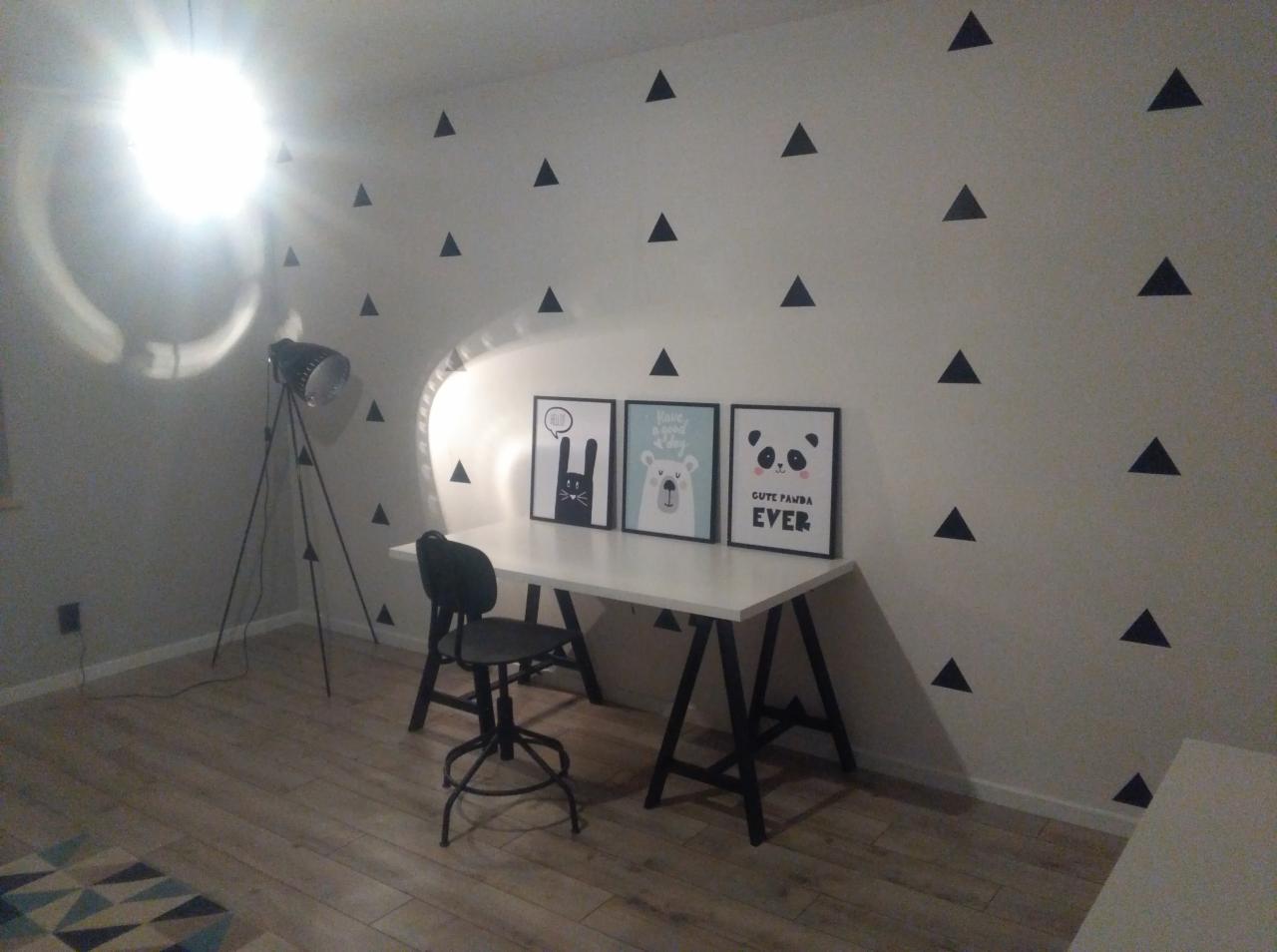 Pokój dziecięcy po remoncie: białe ściany z czarnymi trójkątami, białe biurko z czarnymi nogami, trzy obrazki w czarnych ramkach, lampa na trójnogu i czarne krzesło obrotowe na tle paneli podłogowych.