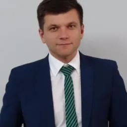 Portret mężczyzny w granatowej marynarce i zielonym krawacie na jasnym tle. Uśmiechnięty, z krótkimi ciemnymi włosami, patrzy wprost w obiektyw. Profesjonalny wizerunek.