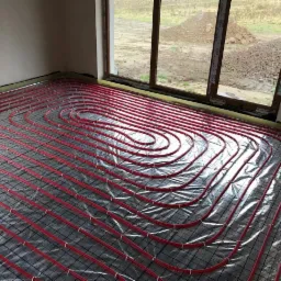 Czerwone rury ogrzewania podłogowego ułożone na srebrnej folii z siatką, widok z góry w surowym pomieszczeniu z oknem na zewnątrz.