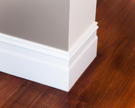 Detal narożnika ściany z białą listwą przypodłogową i parkietem z ciemnego drewna, widoczne oświetlenie górne.