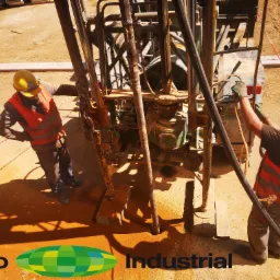 Dwóch pracowników w kamizelkach i kaskach obsługuje maszynę wiertniczą na placu budowy, z widocznymi narzędziami i elementami konstrukcyjnymi.