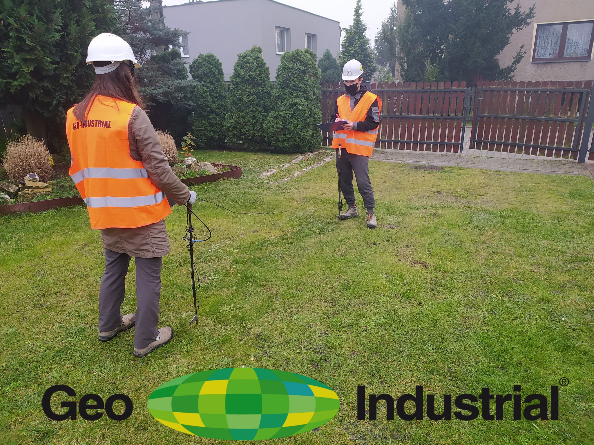 Dwóch inżynierów w kamizelkach odblaskowych i kaskach, jeden z tabletem, drugi z urządzeniem pomiarowym, wykonuje pomiary geodezyjne na trawniku przed domem.
