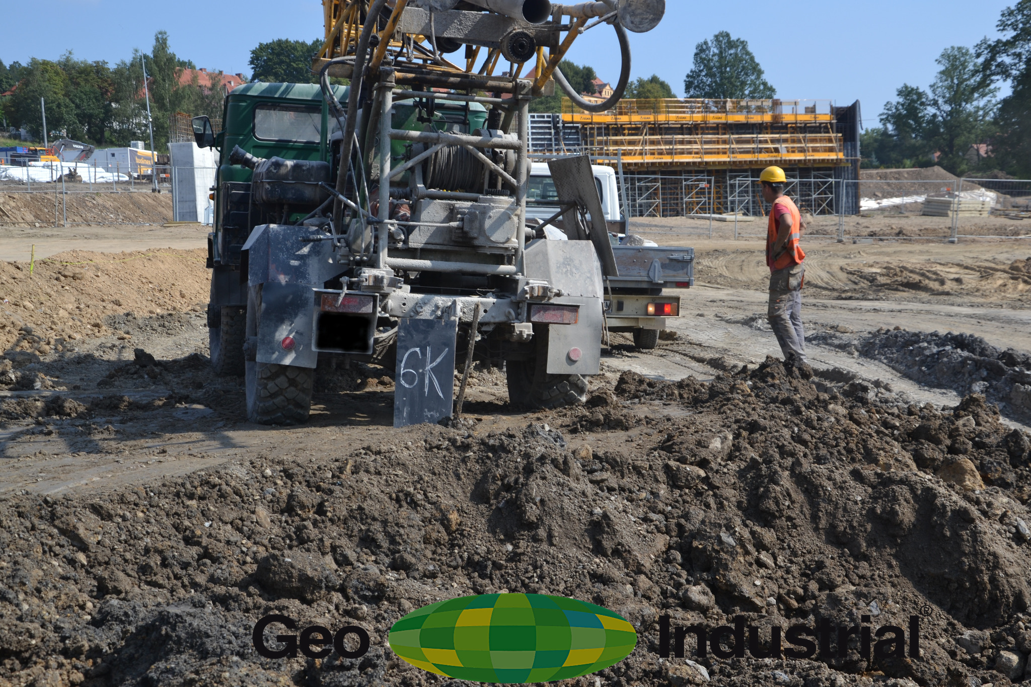Wiertnica zamontowana na ciężarówce na placu budowy, pracownik w kasku i kamizelce odblaskowej stoi obok wykopu, w tle widoczne szalunki.