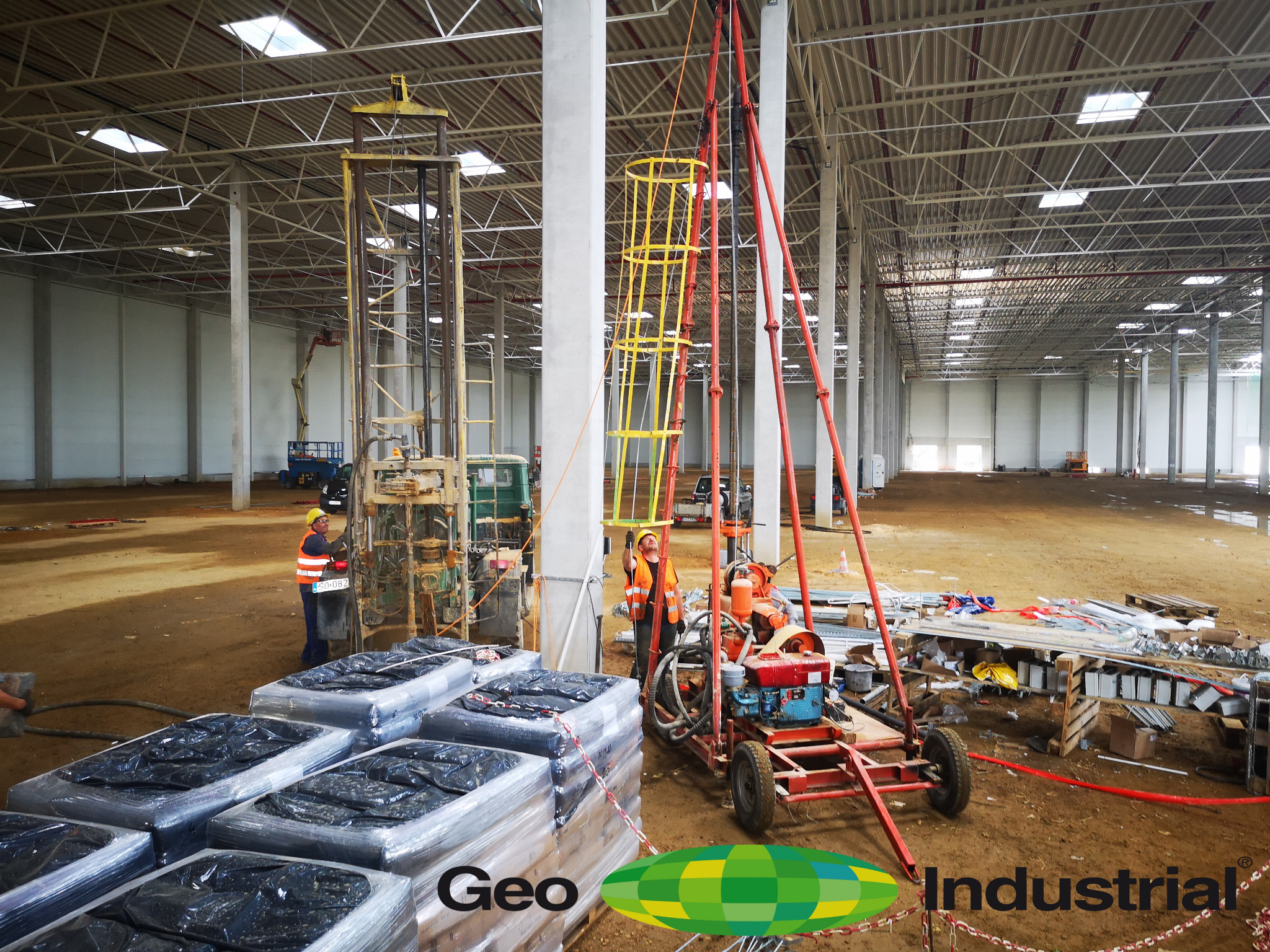 Prace geologiczne w hali magazynowej: dwóch pracowników w kamizelkach odblaskowych obsługuje wiertnice geologiczne, a w tle widoczne są materiały budowlane i konstrukcja hali.