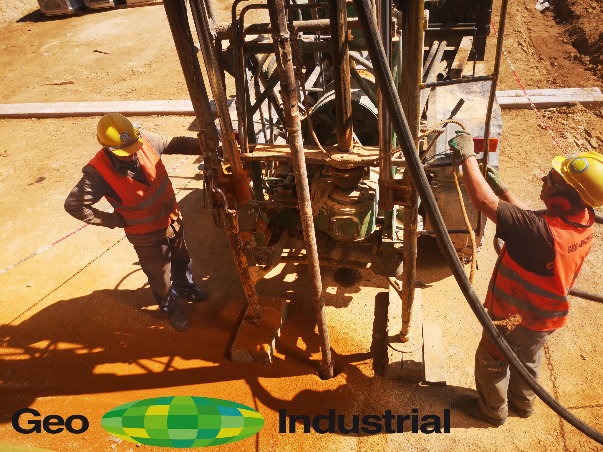 Dwóch pracowników w kamizelkach i kaskach obsługuje maszynę wiertniczą na placu budowy, z widocznymi narzędziami i elementami konstrukcyjnymi.