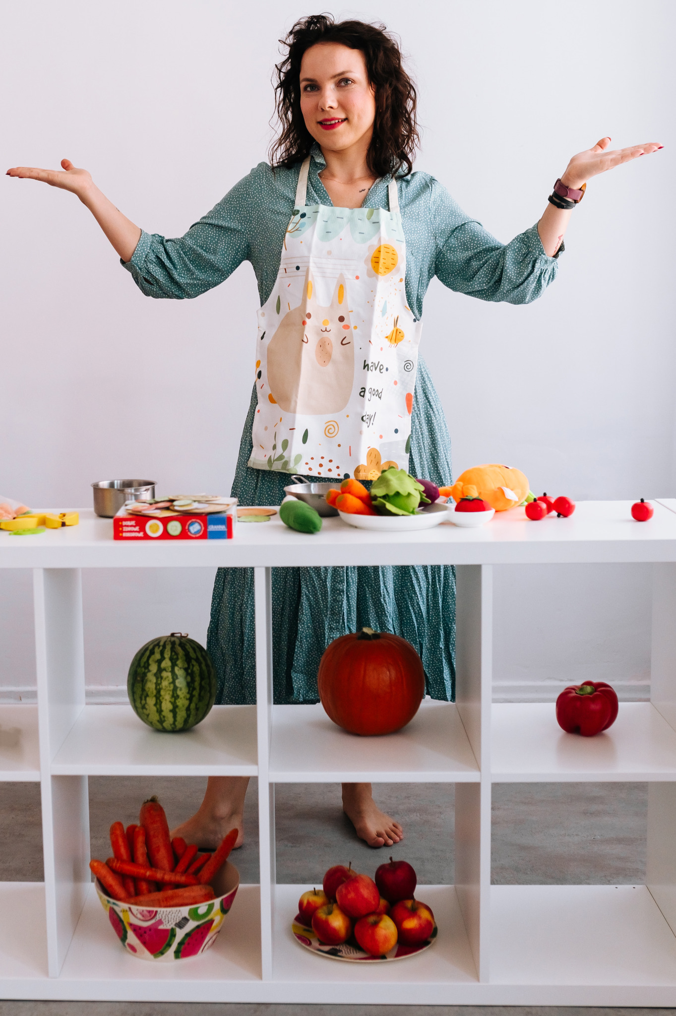 Kobieta w fartuchu z motywem zwierzęcym, prezentująca różnorodne warzywa i owoce na białym regale, z rozłożonymi rękami.